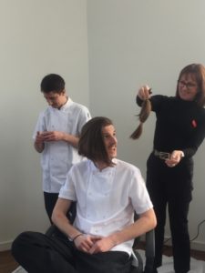 A smiling man who has had his pony tail cut off by woman hairdresser who is holding it