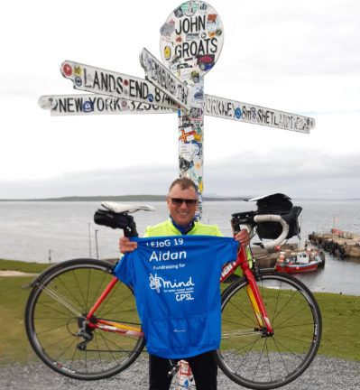Aidan Joy holding his bike with a blue CPSL Mind tshirt in front of the John O Groats sign