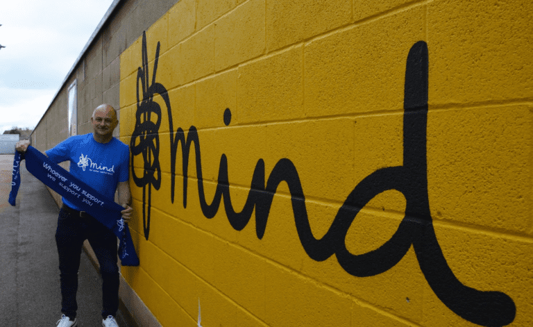 Man in a blue CPSL Mind t shirt holding a whoever you support we support you scarf in front of a yellow wall with the Mind logo