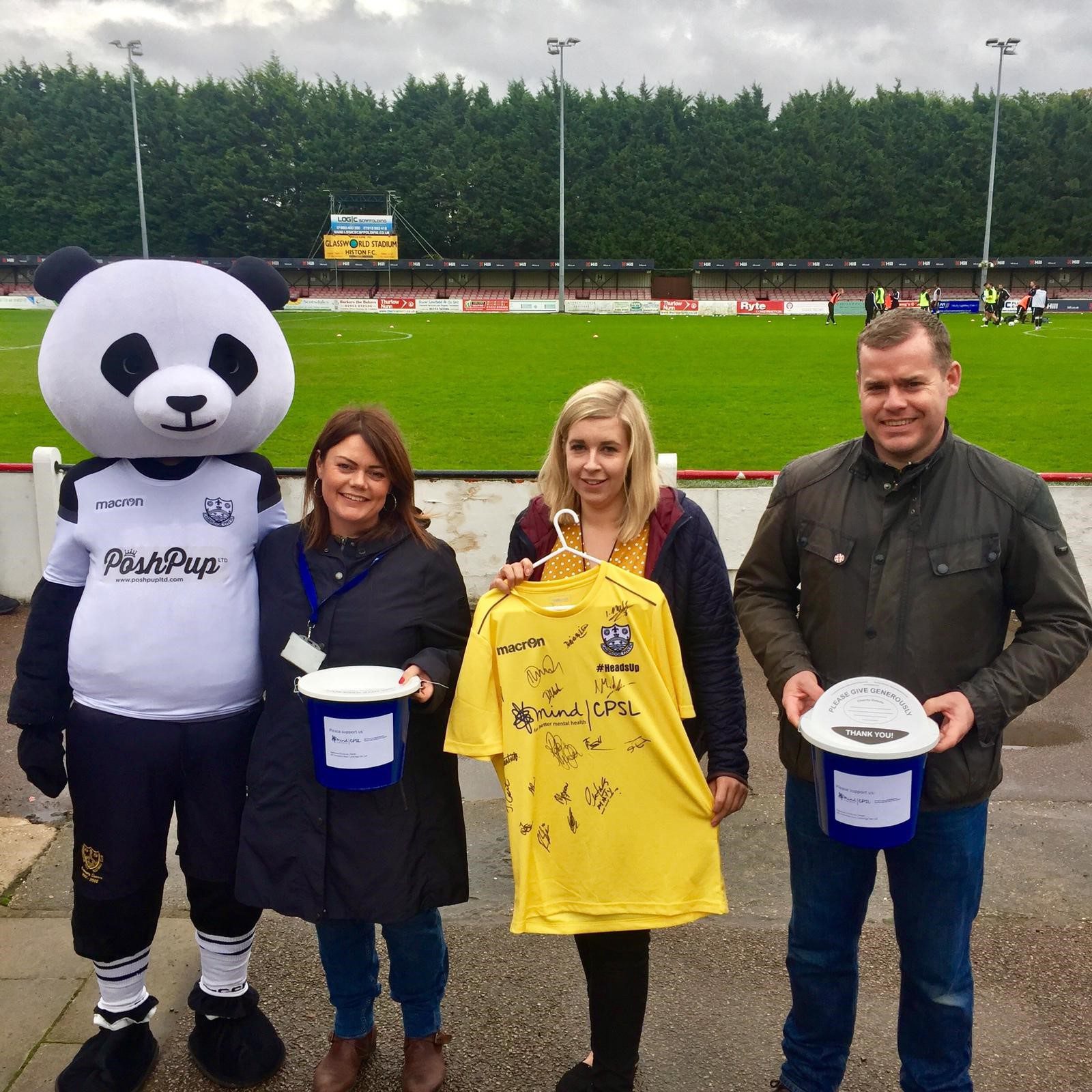 Two women and a man with a Panda mascot, signed football shirt and collection buckets in front of a football pitch