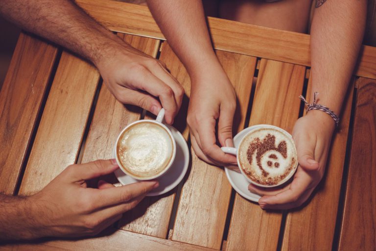 A wooden table with two people's hands holding coffee cups one has a smiley face design in the milk