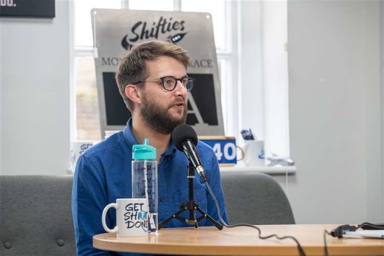 Man with glasses and a beard sat down speaking into a microphone