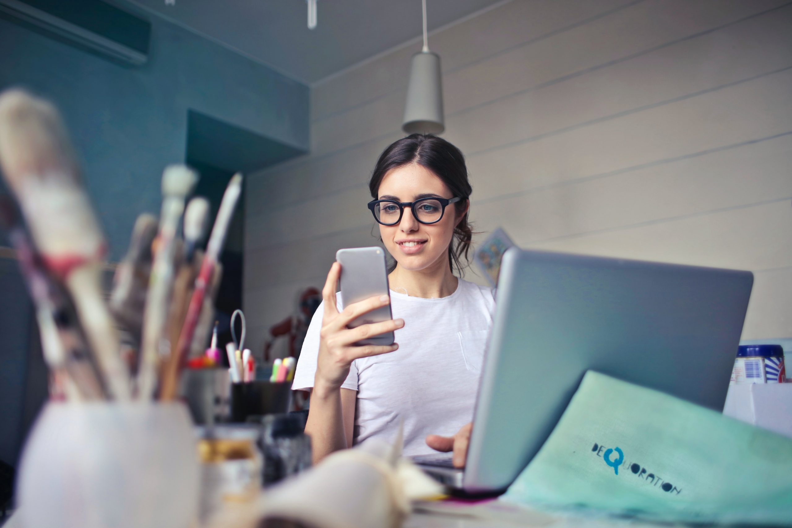 dark haired girl with glasses sat at a desk with her laptop, looking at her phone