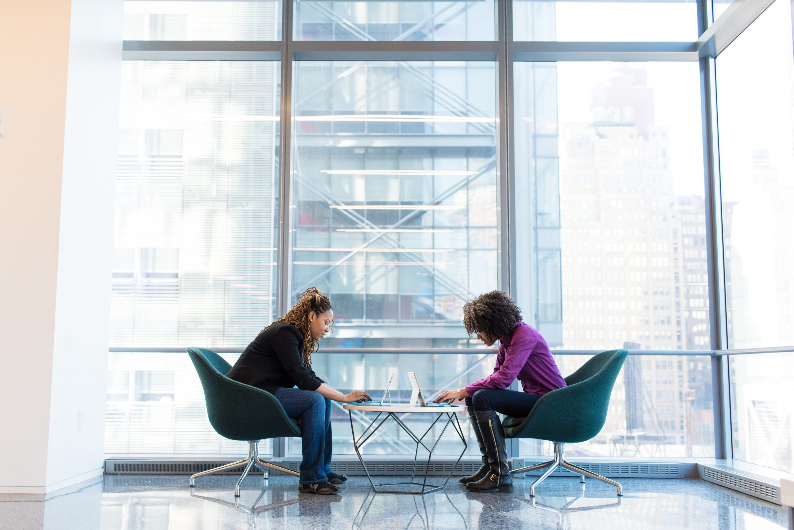 Two women in office setting leaning over small table on laptops - managing mental health in the workplace