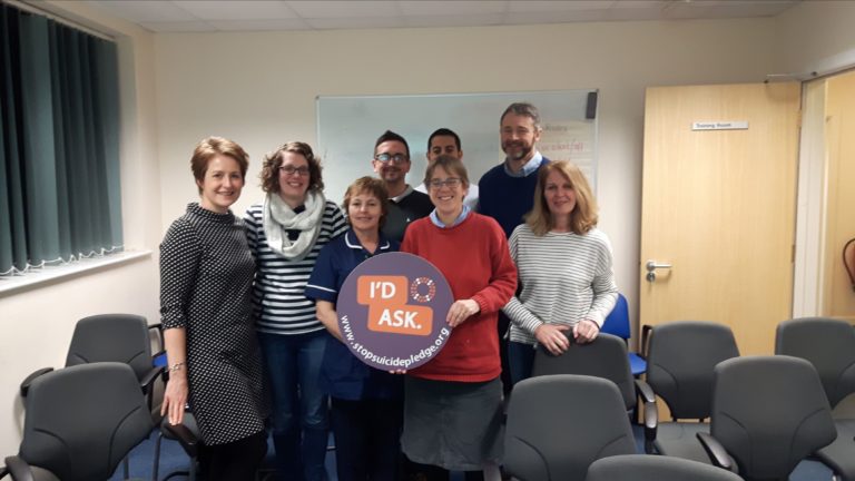 Group of people in a room with grey chairs holding a purple and orange I'd Ask Stop Suicide pledge sign