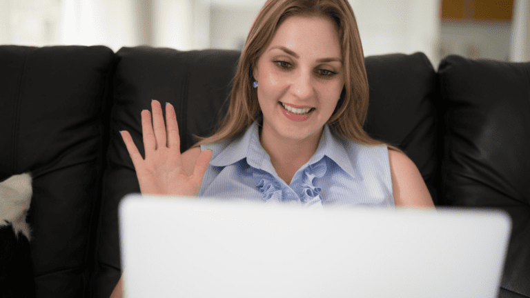 White girl sat on a black sofa waving at her laptop