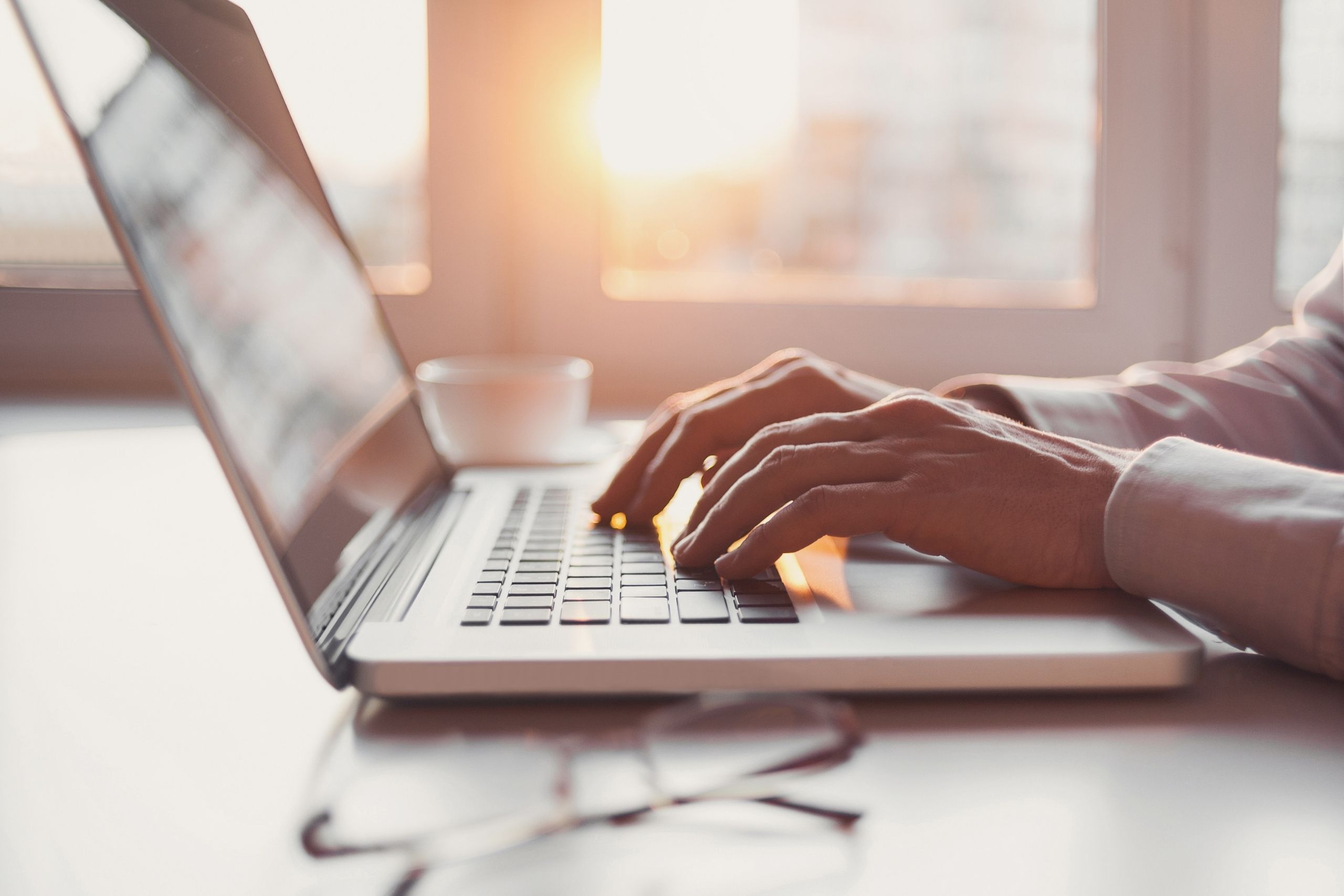 Person in cream jumper typing on laptop. Coffee cup and glasses in background with sun beaming in from windows