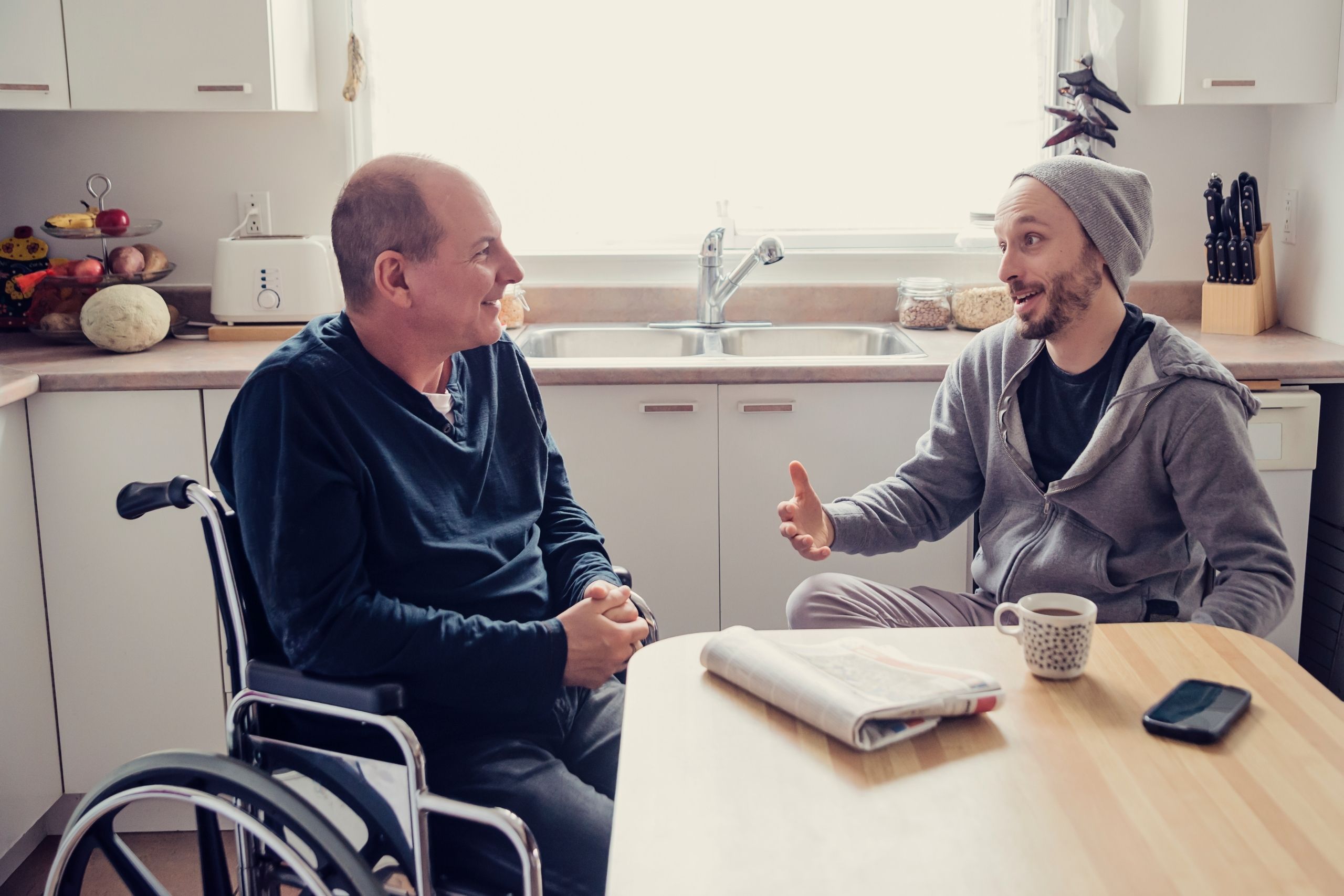 Man in wheelchair speaking to another man sat at a table in a kitchen