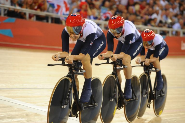 3 Great Britain athletes on bikes with red helmets in a velodrome