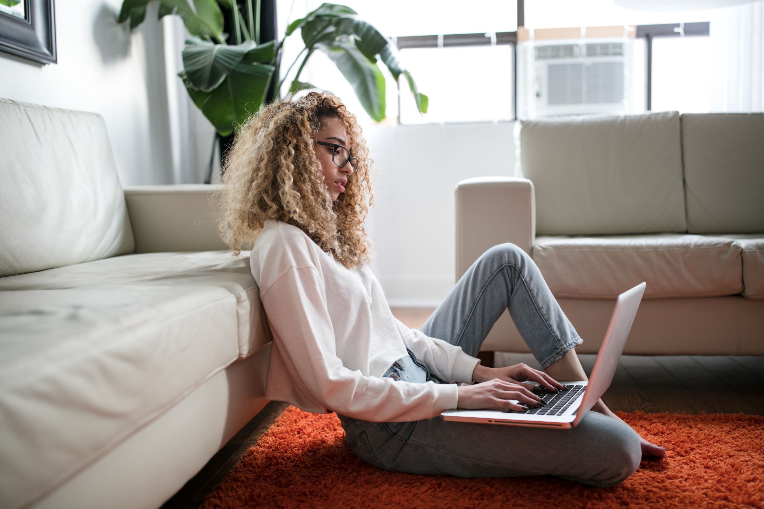 Woman sat on floor using laptop with back against sofa