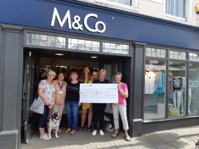 A group of women holding a cheque in front of an M&Co shop