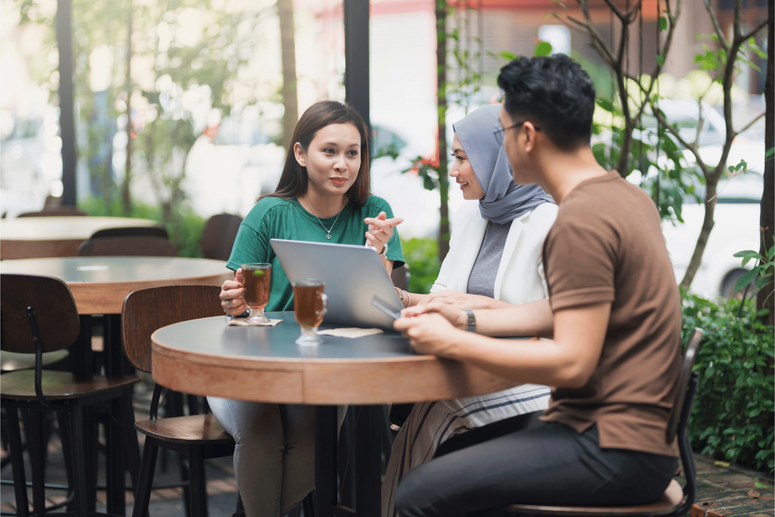 A woman in a green top and long brown hair, a woman with white top and blue hijab and a male wearing a brown top sitting a coffee shop outside with a laptop