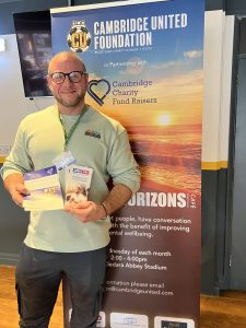 man standing with leaflets in front of Cambridge FC banner 