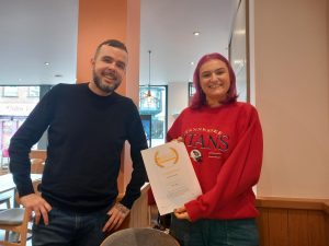Image of man in black top with girl in red jumper holding a certificate