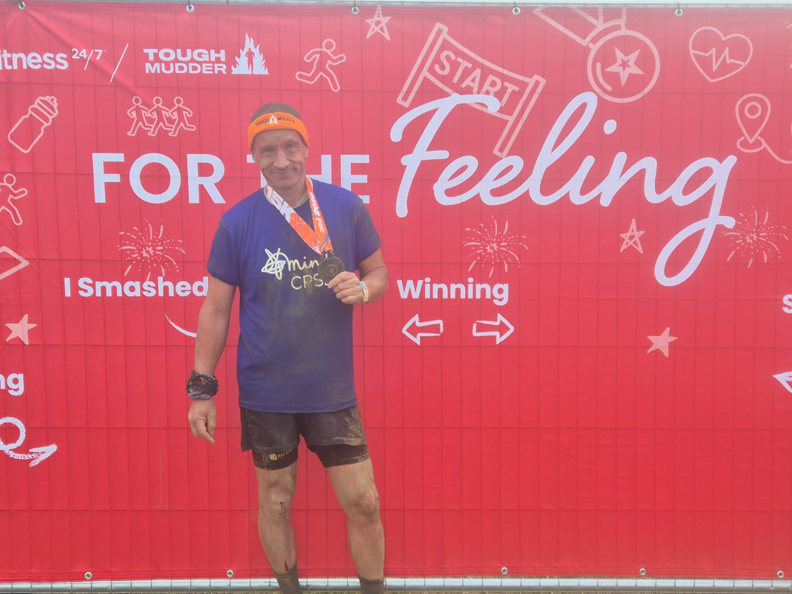 A smiling man covered in mud holding his finisher medal in front of a red Tough Mudder banner.
