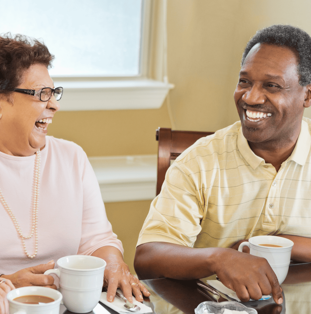 An older couple sharing a laugh together over coffee at a dining table.