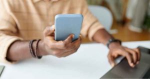 A person sits at a desk using a laptop while holding a smartphone, suggesting multitasking or working online.
