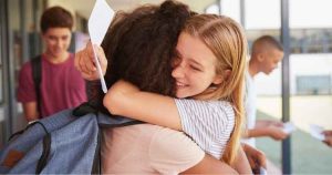 Two students hug outside a school building while holding papers, with other students chatting in the background.