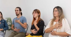 Three people sit with eyes closed, hands on chest and abdomen, practicing a calm breathing exercise.