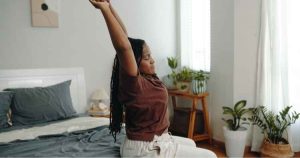 A person sits on the edge of a bed stretching their arms upward in a bright, plant‑filled bedroom.